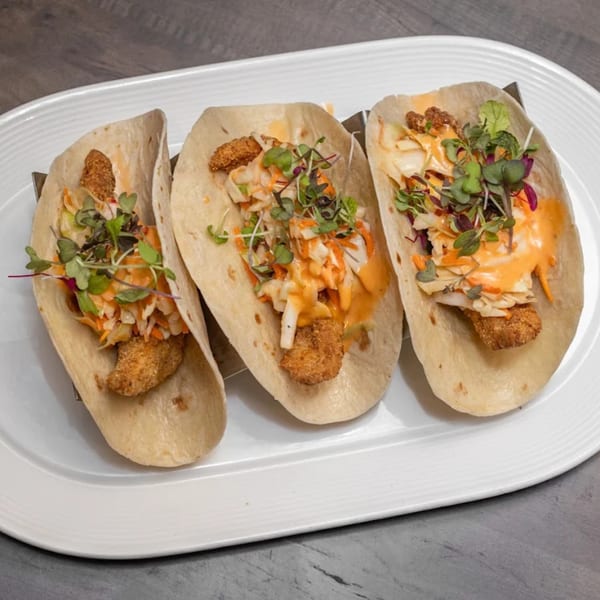 Three American-style tacos with crispy chicken, coleslaw, orange sauce, and microgreens on a white plate.