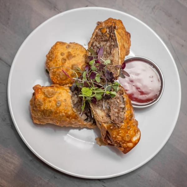 Crispy American egg rolls, one cut open to show the savory filling, garnished with microgreens and served with dipping sauce.